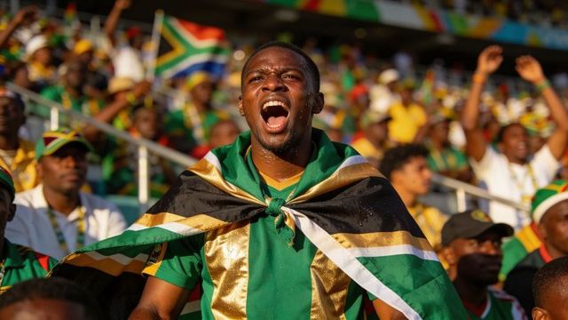 Black supporter shouting draped in national flag leading chant in sunlit stadium crowd