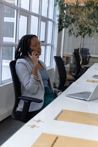 African american woman talking on smartphone at modern open-plan workspace with laptop
