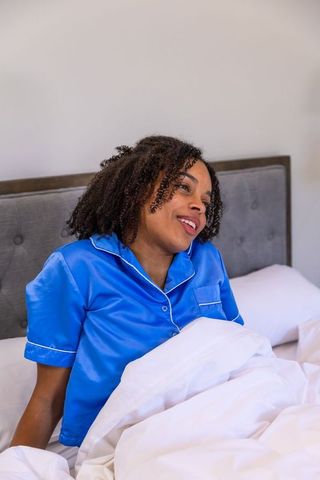 African american woman sitting up in bed wearing blue satin pajamas looking relaxed cozy morning