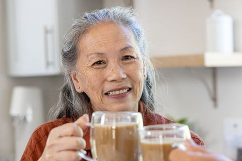 Elderly woman smiling, raising latte mugs with friend, sharing cozy kitchen companionship