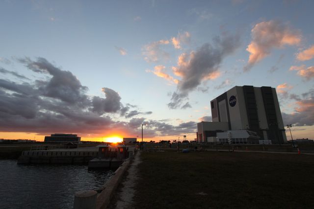This scenic capture of the NASA Kennedy Space Center showcases a beautiful sunset over the Launch Complex 39 area. The illuminated clouds add depth to the evening sky, while prominent structures like the Vehicle Assembly Building, Operations Support Building I and II stand out. Ideal for use in articles, educational materials, and presentations related to space exploration, technology infrastructure, or NASA landmarks.