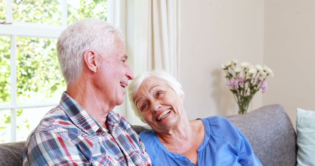 A happy senior couple is enjoying quality time together at home, sitting on a cozy sofa. They are smiling at each other, creating a warm and heartfelt moment perfect for depicting leisure and companionship in senior years. Ideal for content about aging, relationships, elderly care choices, senior lifestyle, and family moments.