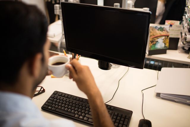Cropped image of businessman holding coffee cup by desktop pc in office