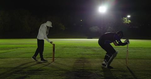 Night cricket under floodlights with umpire adjusting stumps and batsman guarding crease