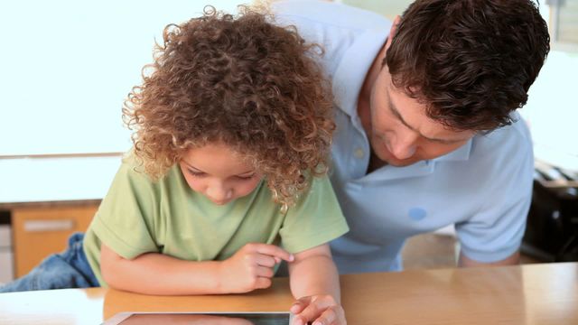 Father and son bonding over shared technological activity using a tablet in a modern kitchen. Ideal for content on family life, parenting, technology in education, and modern family dynamics.