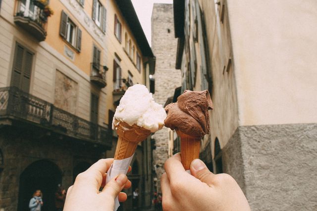 Two individuals are holding delicious ice cream cones, one vanilla and one chocolate, in an urban setting with historic buildings in the background. Suitable for travel blogs, food-related articles, and advertisements showcasing city travel during summer. Can also be used for promoting desserts or exploring European cities.