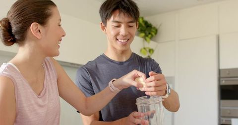 Asian couple preparing banana smoothie in bright modern kitchen, smiling together