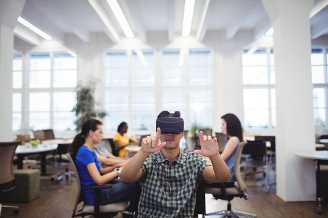 Man wearing virtual reality headset, interacting through hand gestures in a contemporary office environment. Colleagues working in background. Ideal for illustrating concepts related to digital innovation, modern workplace, and virtual reality technology.