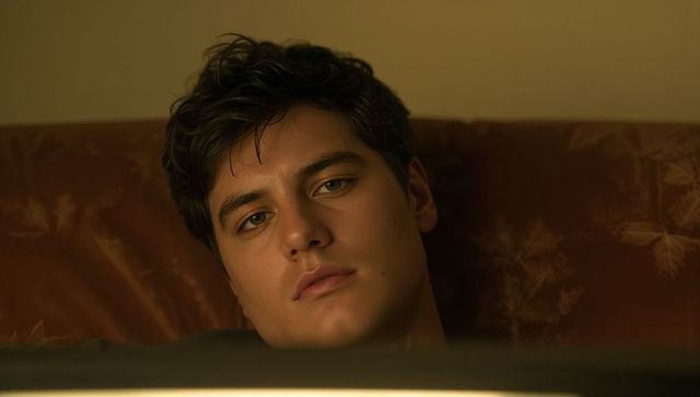 Reclining young man gazing downward in warm screen light on brown sofa, introspective portrait