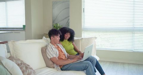 Young asian man and black woman browsing laptop together on cozy living room sofa