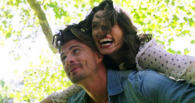 A young Caucasian couple enjoys a playful moment outdoors, with the woman piggybacking on the man, both smiling and surrounded by greenery, with copy space. Their joyful expressions and the sunlit backdrop suggest a carefree, romantic day in nature.