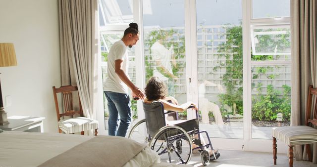 Biracial woman in wheelchair and male partner looking out of window in sunny bedroom. wellbeing and domestic lifestyle with physical disability.