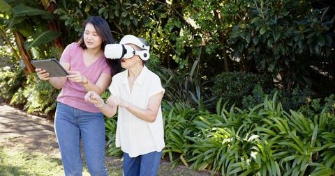 Asian caregiver guiding senior woman using vr headset in sunlit garden for elder wellness