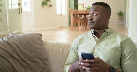 African american man holding smartphone glancing sideways on beige sofa in sunlit modern home