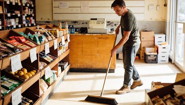 Local market worker sweeping floor with broom among shelves and crates during cleanup