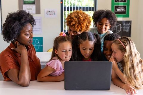 African American teacher guiding diverse elementary students around laptop for teamwork