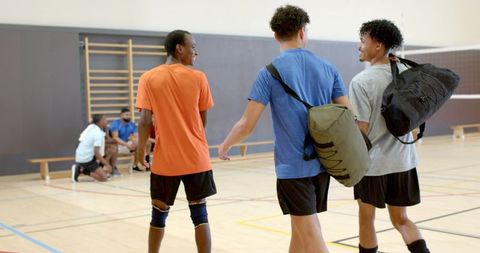Diverse male athletes walking across gym carrying duffel bags after volleyball practice