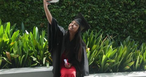 Indian graduate taking selfie in cap and gown on sunlit campus courtyard holding diploma