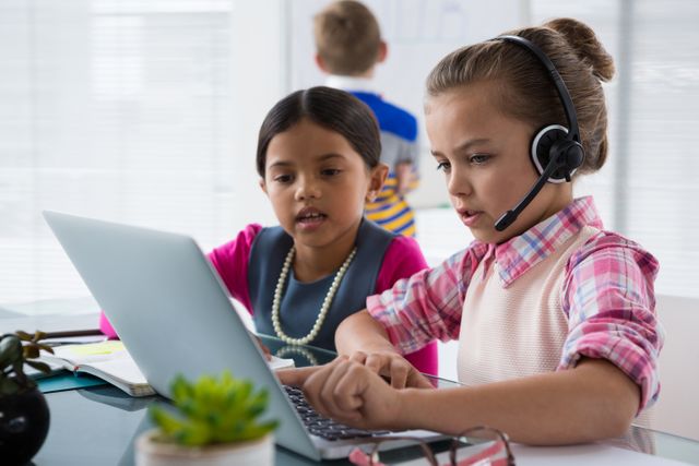 Children working together in an office environment, using a laptop and headset. Ideal for concepts related to teamwork, early education, young professionals, and technology in learning. Suitable for educational materials, business training programs, and advertisements promoting collaborative learning and communication skills.