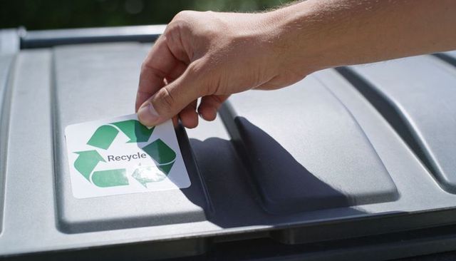 Hand applying recycle sticker on gray outdoor bin lid sunlit ecofriendly recycling action