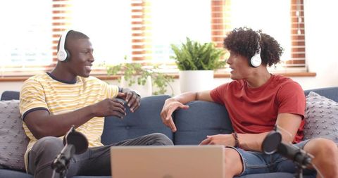 Two men recording podcast on sofa wearing headphones in home studio with mics and laptop