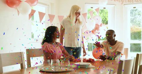 Friends celebrating at sunlit dining table with pastel balloons and confetti