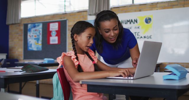 Diverse smiling female teacher helping schoolgirl sitting in classroom, using laptop. children in primary school during.