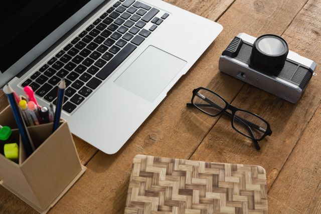Close-up of laptop, camera, spectacles, notepad and pencil holder on wooden plank