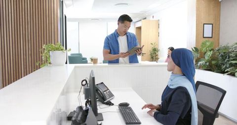 Receptionist in navy hijab assisting visitor with tablet at modern bright office reception