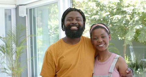 Smiling african father and daughter standing arm in arm by sunlit glass doors at home