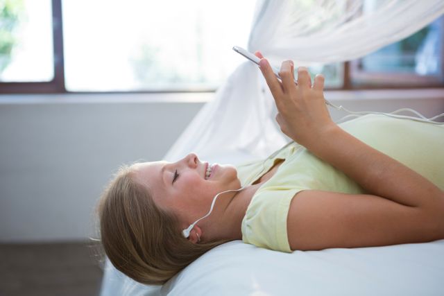 Side view of smiling girl listening music while using smartphone on bed at home