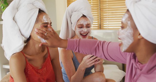 Diverse group of happy female friends with towels on heads and cleansing masks taking selfie at home. female friends hanging out enjoying leisure time together.