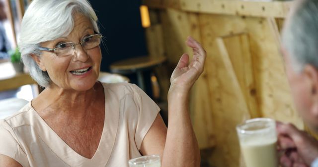 Senior woman enjoying a conversation with a friend in a cozy cafe. She is smiling and gesturing with her hand while holding a drink. Use this for topics on social connections in older age, coffee shop culture, and elderly lifestyles.