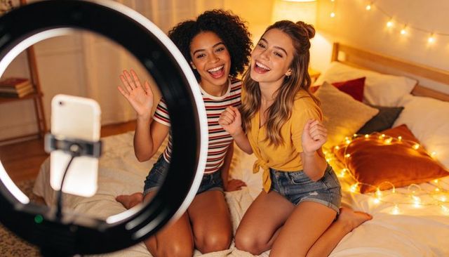 Two friends recording vertical video on bed with ring light, smiling and waving in cozy bedroom