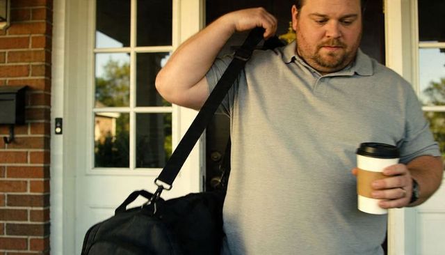 Commuter leaving home carrying duffel and holding coffee on suburban porch doorway