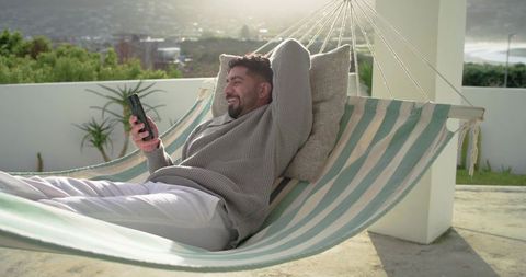 Indian man relaxing on striped hammock with phone on sunny rooftop terrace coastal view