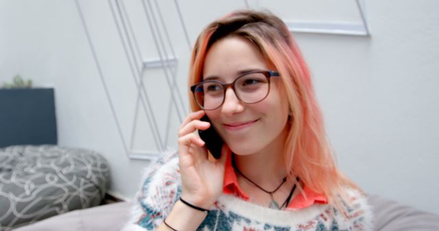 Young woman with colorful hair wearing glasses is smiling while talking on mobile phone indoors. She is dressed casually and looks comfortable. Perfect for illustrating modern communication, casual conversations, youth culture or customer service scenarios.