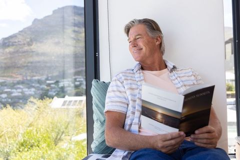 Mature man relaxing by window reading sunset magazine with hillside view, sunlit interior