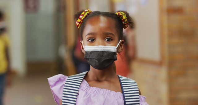 Portrait of african american schoolgirl wearing face mask, standing in corridor looking at camera. children in primary school during coronavirus covid 19 pandemic.