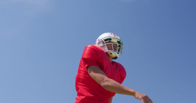 Football player in bright red jersey and helmet practicing on grass field against clear blue sky. Perfect for marketing of sports equipment, team events, health and fitness campaigns or inspirational posters.