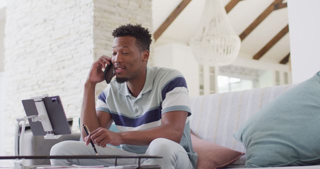 Young man is multitasking by using a tablet and smartphone while sitting in a home office setting. This image is great for showcasing remote work, telecommuting, technology use, and casual work environments. Suitable for website banners, blog posts on remote work, and advertisements for technology or remote working tools.