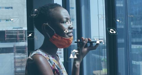 Masked woman giving voice command indoors by window with eco icons