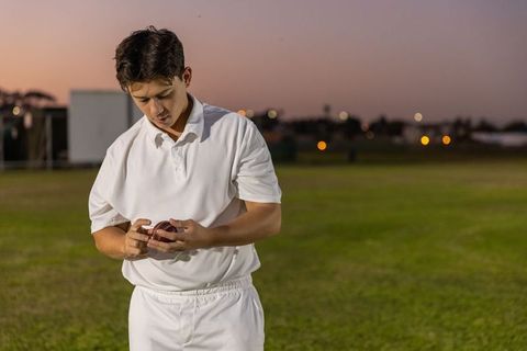 Young cricketer standing on dusk outfield inspecting red leather ball wearing white uniform