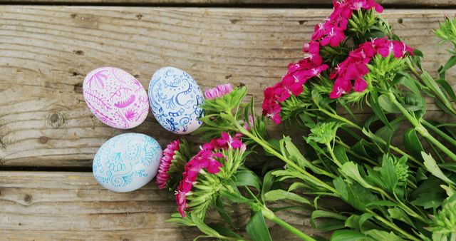 Decorated Easter eggs lie next to vibrant pink flowers on a rustic wooden background, with copy space. Easter eggs and spring flowers symbolize rebirth and new beginnings, marking the festive spirit of the holiday.