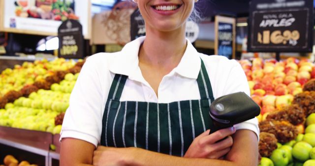Smiling female grocery store worker wearing striped apron holding barcode scanner in front of fruit display. Ideal for use in content related to retail employment, customer service, grocery store operations, and demonstrating positive employee attitude.