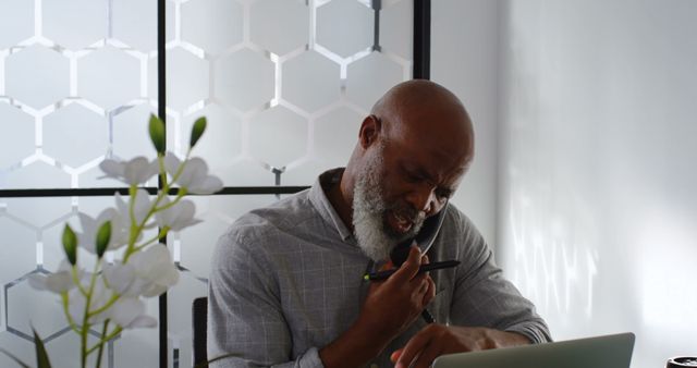 Senior man concentrating while multitasking, working remotely using laptop and phone, showcasing modern remote work environment with decorative flowers. The image can be used for articles or promotions related to home office setup, productivity tips, remote work, senior professionals adapting to technology, and multitasking strategies.