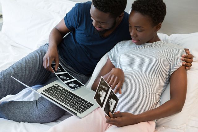 Expecting couple sitting on bed, viewing sonogram images on laptop, sharing a tender moment. Ideal for use in articles or advertisements related to pregnancy, family bonding, parenthood, and home life. Perfect for illustrating themes of love, anticipation, and modern technology in family planning.