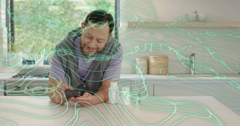 Older asian man leaning on kitchen counter holding smartphone and gadget with teal lines