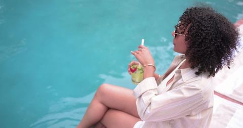 African-american woman relaxing poolside sipping cocktail in sunglasses, summer lifestyle