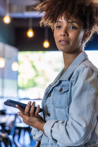 African american woman holding smartphone in cozy coffee shop, denim jacket portrait, natural light